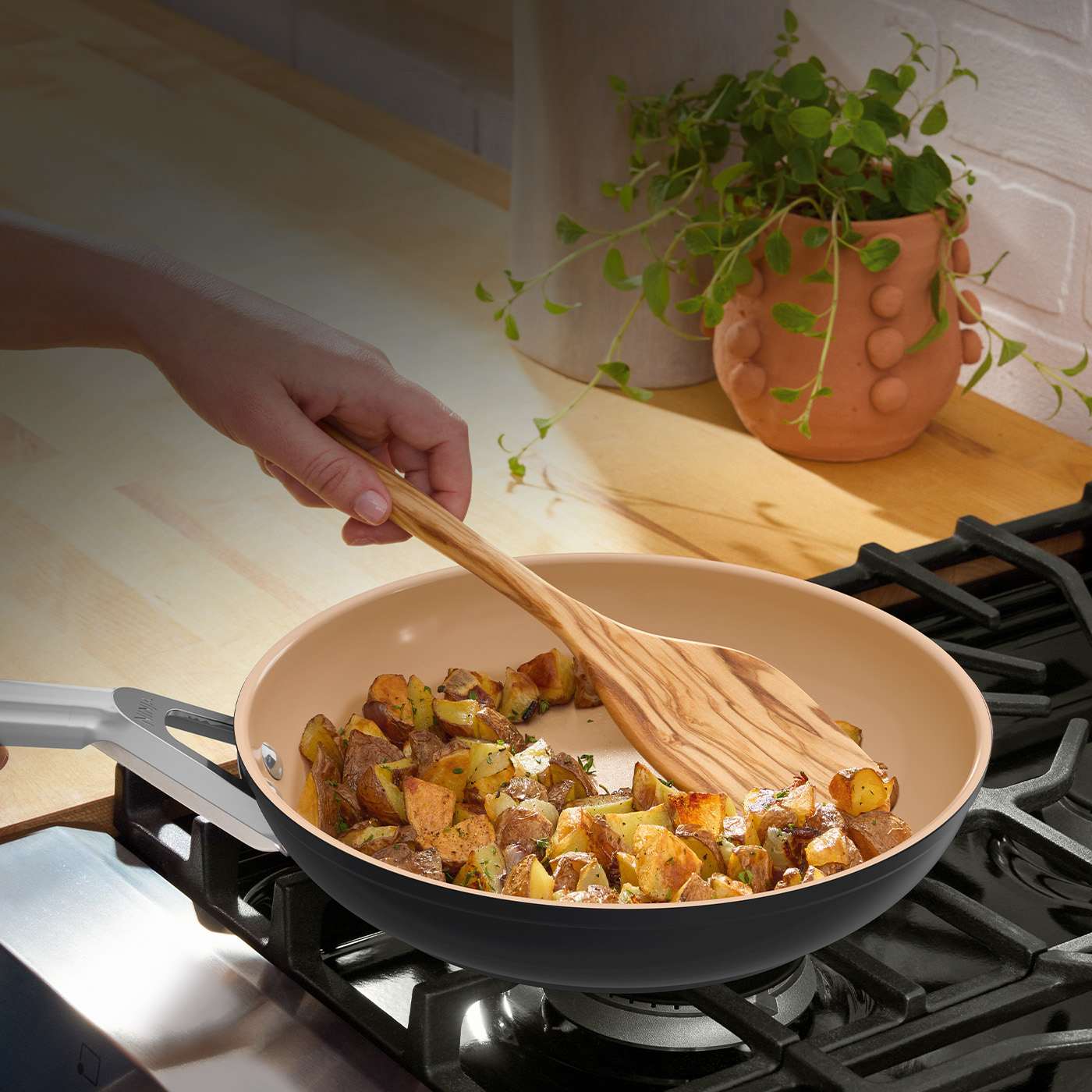 Hand stirring potatoes in a terracotta and grey ceramic Ninja frying pan on a gas hob