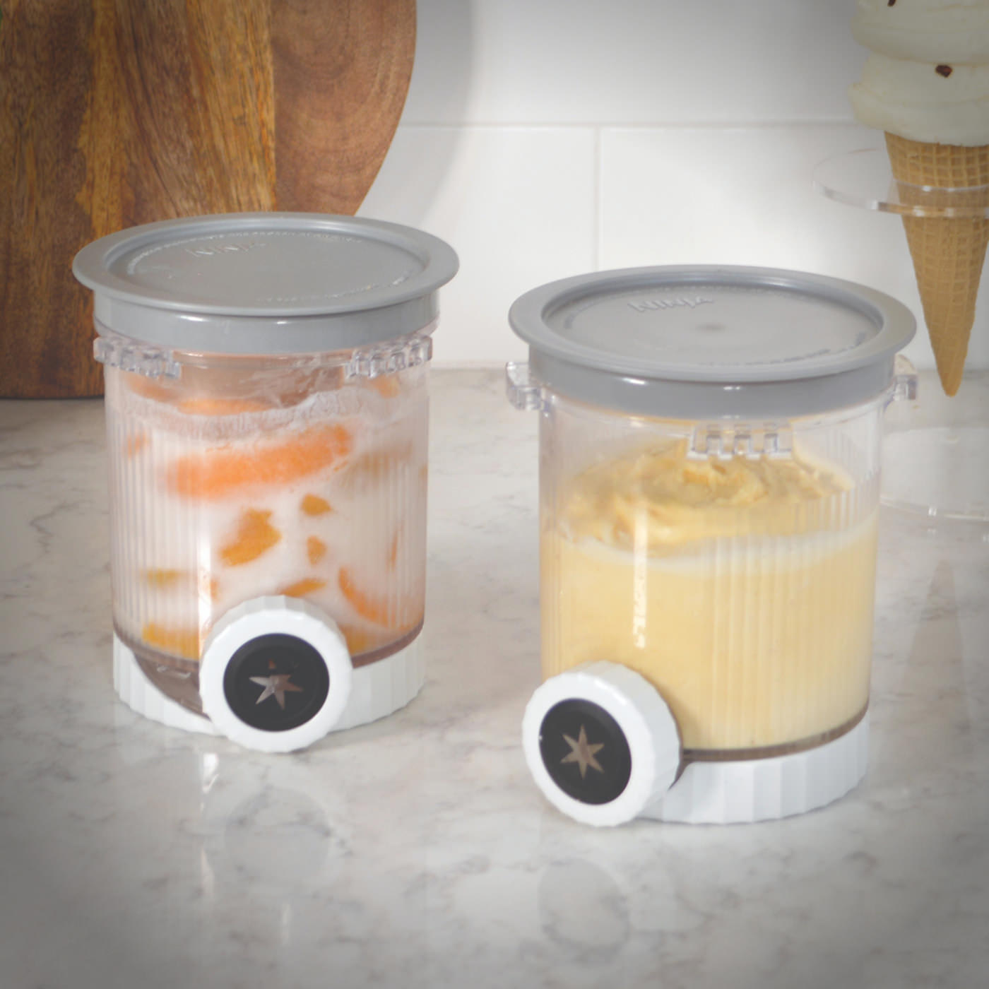 Two Ninja CREAMi dessert containers filled with swirled frozen treats, sitting on a marble countertop with visible nozzles attached.